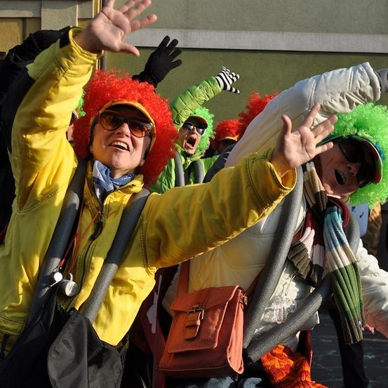 Eine Gruppe von Menschen in gelben und grünen Kostümen mit roten und grünen Perücken jubelt und hebt ihre Hände in der Luft.