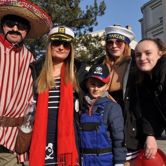 Eine Gruppe von Menschen in verschiedenen Kostümen, darunter ein Matrose, eine BMW-Kappe, ein Mexikaner und zwei Frauen, posiert für ein Foto. Im Hintergrund sind ein Gebäude und Bäume zu sehen.