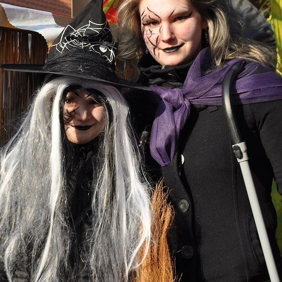 Zwei Frauen in Halloween-Kostümen, eine als Hexe und die andere als Hexenbegleiterin, stehen gemeinsam vor einer Kulisse.