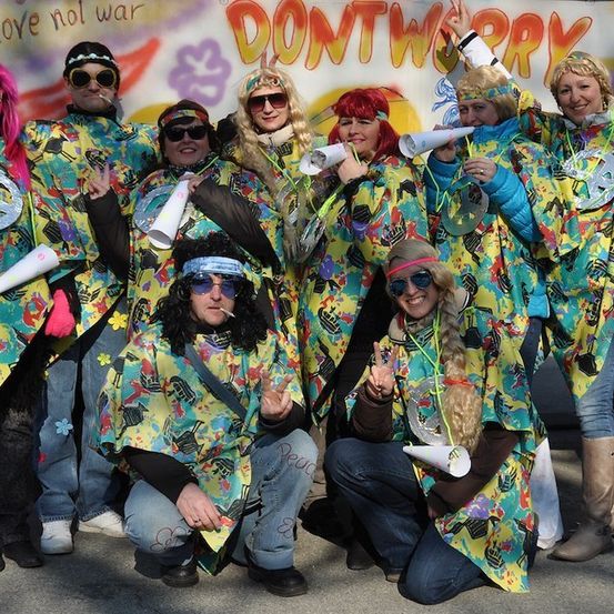 Eine Gruppe von Menschen in bunten, bunt gestreiften Outfits posiert für ein Foto mit Friedenszeichen und Zigaretten vor einem Banner, auf dem 'DONT WORRY' und 'LOVE NOT WAR' steht.