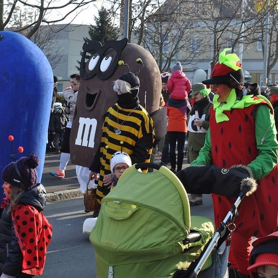 Eine Gruppe von Menschen in Kostümen, darunter ein großer blauer Ballon, eine Biene und eine Erdbeere, gehen auf einer Straße mit einem Kinderwagen in der Mitte.