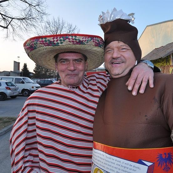 Zwei Männer in Kostümen, einer trägt einen Sombrero und der andere ein braunes Kostüm mit Hut, stehen lächelnd auf einem Parkplatz.