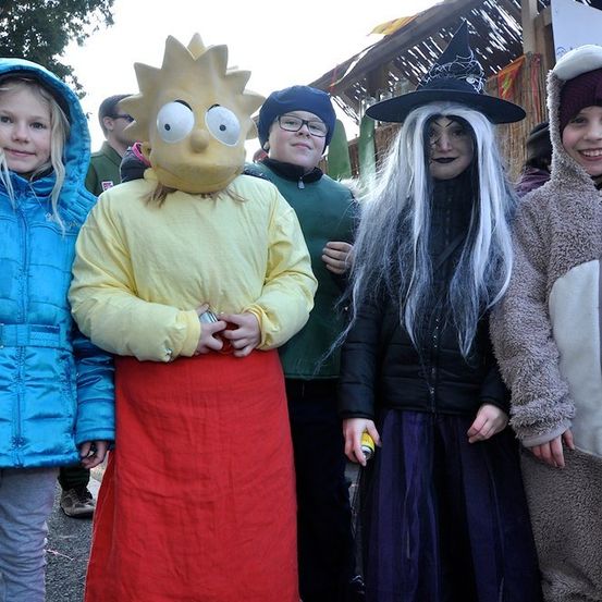 Eine Gruppe von Kindern in verschiedenen Kostümen, eines davon mit einer Marge-Simpson-Maske, steht vor einer Halloween-Dekoration.