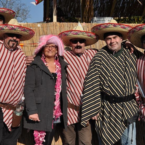 Fünf Personen in mexikanischen Kostümen, einschließlich Sombreros, stehen vor einer Bambuswand. Eine Frau in der Mitte trägt einen rosa Hut und eine Brille.