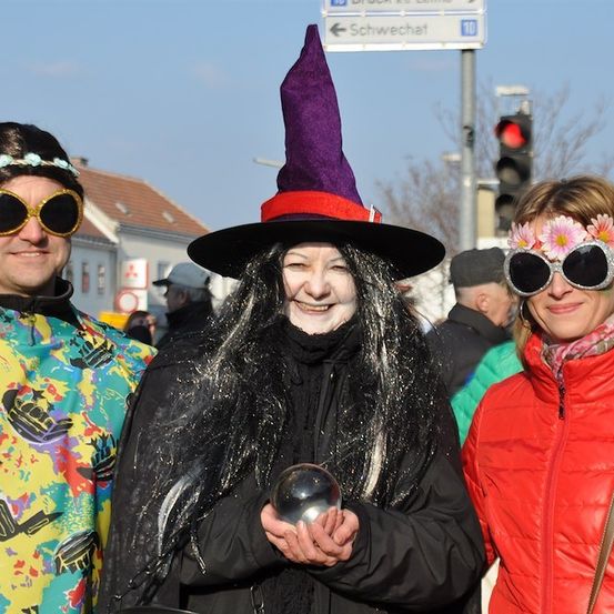 Drei Menschen in Kostümen stehen zusammen, eine Frau mit Hexenhut und Kristallkugel, eine andere mit Blumenkopfband und Sonnenbrille und ein Mann mit buntem Shirt und Sonnenbrille.