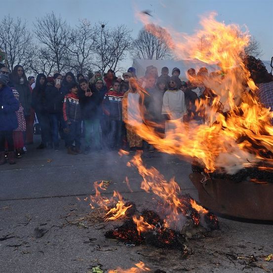 Eine Menschenmenge, darunter Kinder, versammelt sich um ein großes Lagerfeuer und beobachtet, wie es mit Flammen hell brennt.