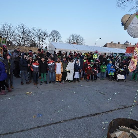 Eine Menschenmenge, darunter Kinder in Kostümen, versammelt sich auf einem Freigelände, möglicherweise für ein Festival. Ein Mensch in einem großen Kostüm steht vor ihnen.