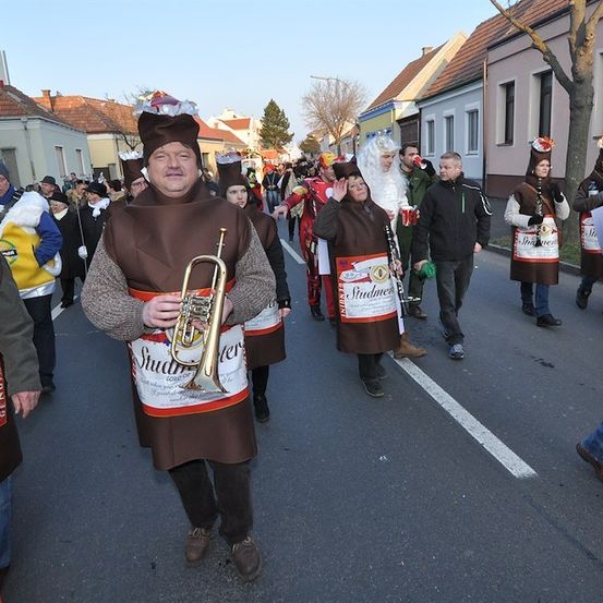 Eine Gruppe von Menschen in Kostümen zieht bei einem Umzug durch die Stadt, einige halten Musikinstrumente. Die Kostüme sind thematisch auf Bierflaschen ausgerichtet, und sie gehen auf einer Straße mit Gebäuden auf beiden Seiten.