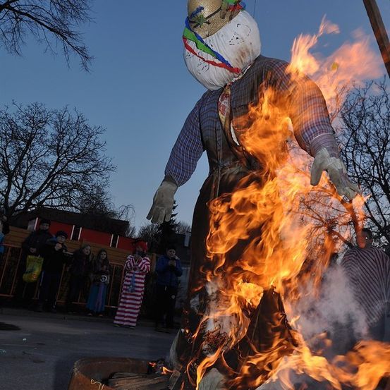 Eine große Strohpuppe brennt in einem Feuerkorb, während Menschen hinter einem Zaun zusehen. Die Puppe ist in einem bunten Outfit und einem Hut gekleidet.