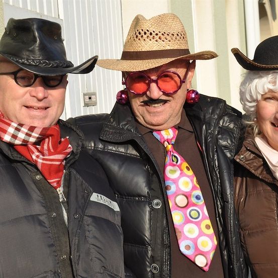 Drei Menschen in Kostümen und Hüten stehen zusammen und lächeln für ein Foto. Der Mann links trägt einen schwarzen Cowboyhut und eine Brille. Der Mann in der Mitte trägt einen Strohhut, eine Sonnenbrille und eine bunte Krawatte. Die Frau rechts trägt einen schwarzen Hut und eine weiße Perücke.