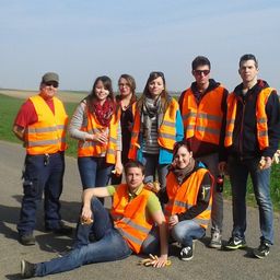 Eine Gruppe von Menschen in Warnwesten steht und sitzt auf einer Straße, lächelt und posiert für ein Foto. Sie befinden sich auf einem Feld mit klarem Himmel.