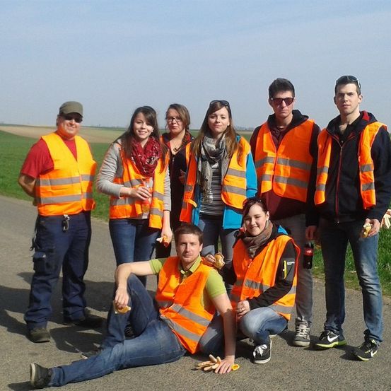 Eine Gruppe von Menschen in Warnwesten steht und sitzt auf einer Straße, lächelt und posiert für ein Foto. Sie befinden sich auf einem Feld mit klarem Himmel.
