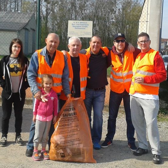 Eine Gruppe von Menschen in orangefarbenen Westen und einem kleinen Mädchen mit einem Müllsack stehen draußen und lächeln für ein Foto. Hinter ihnen befindet sich ein Schild und ein Zaun.