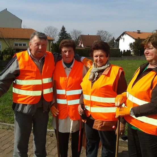 Vier Erwachsene in orangefarbenen Warnwesten stehen vor einem Rasen mit Häusern im Hintergrund.