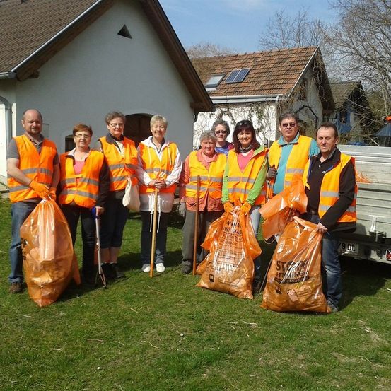 Eine Gruppe von Freiwilligen in orangefarbenen Westen und Handschuhen, die orange Müllsäcke halten, steht vor einem Haus mit Solarmodulen auf dem Dach.