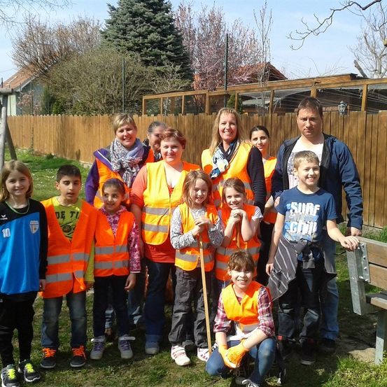 Eine Gruppe von Kindern und Erwachsenen in Warnwesten posiert für ein Foto in einem Garten mit einem Holzzaun und einem Haus im Hintergrund.