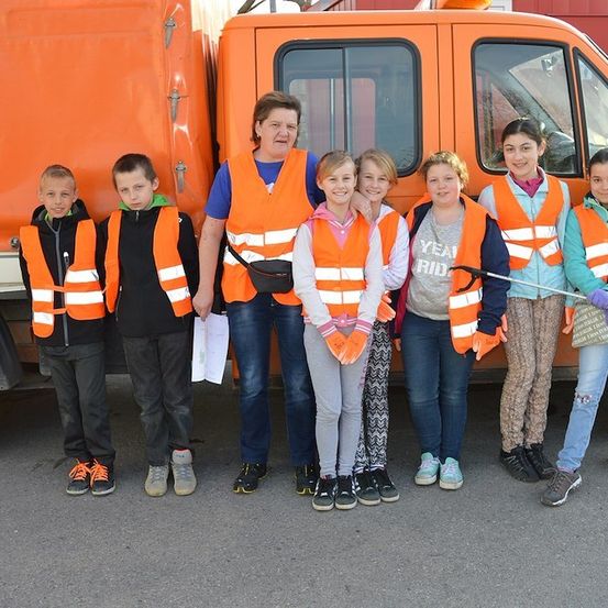 Eine Gruppe von Kindern und einer erwachsenen Frau in orangefarbenen Warnwesten vor einem orangefarbenen Lastwagen. Die Frau hält ein Papier und die Kinder lächeln.