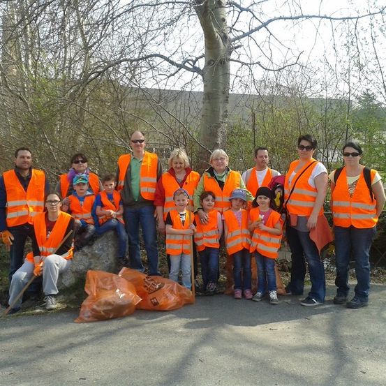 Eine Gruppe von Menschen, einschließlich Erwachsener und Kindern, die orange Westen tragen und mit Müllsäcken in einem Park für ein Foto posieren.