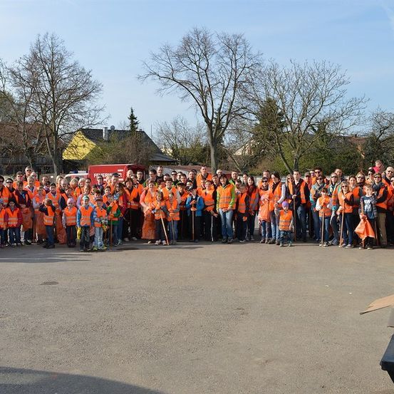Eine Gruppe von Menschen in orangefarbenen Westen und Mützen steht in einer Reihe, wahrscheinlich für eine Gemeinschaftsdienstveranstaltung. Bäume und Gebäude sind im Hintergrund.