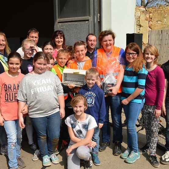 Eine Gruppe von Kindern und Erwachsenen steht zusammen draußen, einige tragen Sicherheitswesten und halten Geschenke. Eine Frau in einer orangefarbenen Weste hält ein verpacktes Geschenk und ein Buch. Hinter ihnen befindet sich eine Ziegelmauer und ein Holzzaun.