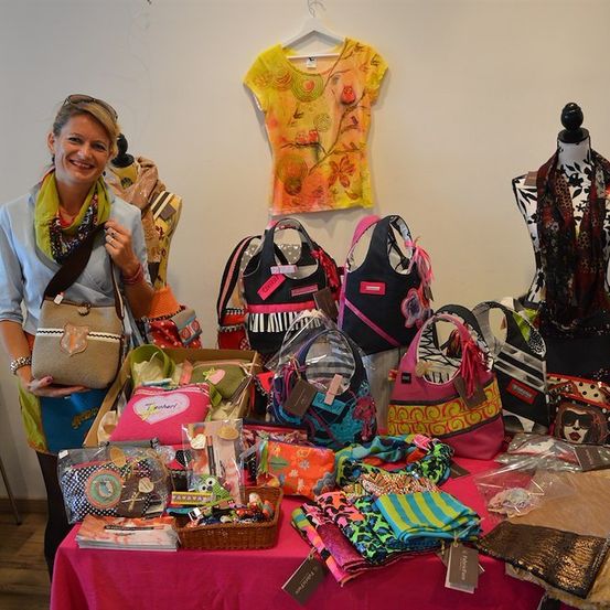 Eine Frau steht hinter einem Tisch voller bunter Taschen, Schals und anderer Accessoires. Sie lächelt und hält eine Tasche. Der Tisch ist mit einem rosa Tischtuch bedeckt.