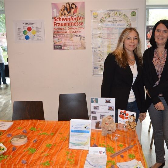 Zwei Frauen stehen hinter einem Tisch mit verschiedenen ausgestellten Gegenständen, darunter ein Plüschtier, eine Kerze und ein Schild, das 'Schwadorfer Frauenmesse' besagt. Der Tisch ist mit einer orangefarbenen Tischdecke bedeckt und es hängen Poster an der Wand hinter ihnen.