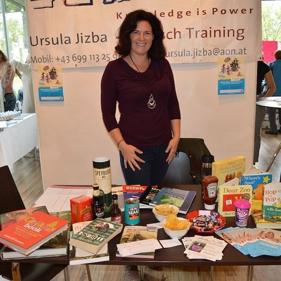 Eine Frau steht hinter einem Tisch mit Büchern, Snacks und Werbematerialien. Sie trägt ein weinrotes Shirt und eine Halskette. Hinter ihr steht ein Banner mit 'Wissen ist Macht' und 'Ursula Jizba' mit einer Kontakt-E-Mail.