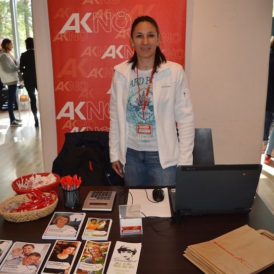 Eine Frau steht hinter einem Schreibtisch mit Laptop, Taschenrechner und verschiedenen Broschüren. Sie trägt eine weiße Jacke und einen Ausweis. Hinter ihr ist ein roter Banner mit dem Text AKNO.