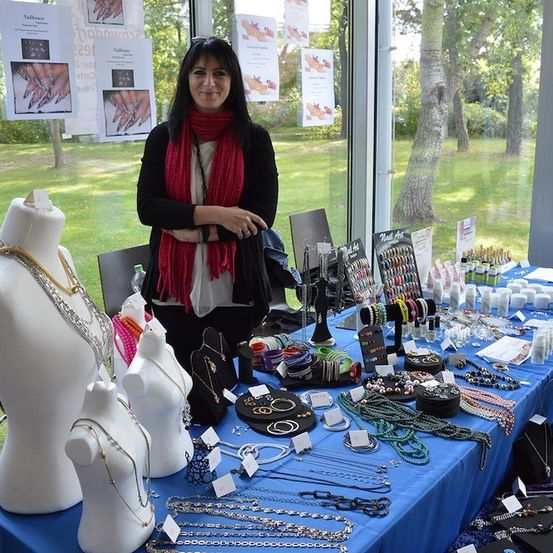 Eine Frau steht hinter einem Tisch voller Schmuck, Mannequins und Nagellack. Der Tisch ist mit einem blauen Tuch bedeckt und vor einem großen Fenster aufgestellt.
