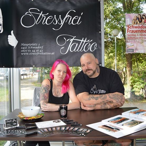 Ein Mann und eine Frau mit Tattoos sitzen an einem Tisch mit verschiedenen Gegenständen, darunter eine Schüssel und ein Ordner, vor einem Banner, das ein Tattoostudio bewirbt.