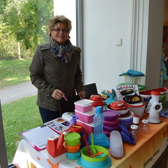 Eine Frau steht hinter einem Tisch, der mit bunten Plastikbehältern und Utensilien gefüllt ist, lächelt und hält einen Stift. Der Tisch ist mit einer gemusterten Tischdecke bedeckt. Im Hintergrund befindet sich ein Rasenbereich und ein Gebäude.