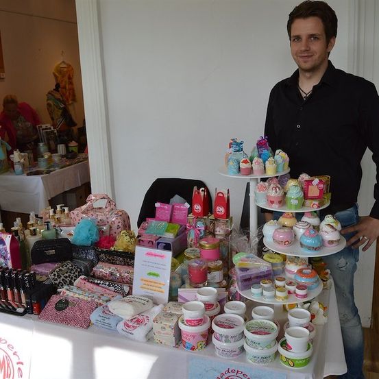 Ein Mann steht hinter einem Tisch voller verschiedener Schönheitsprodukte, darunter Cupcakes, Lotionen und Make-up. Er trägt ein schwarzes Hemd und Jeans.