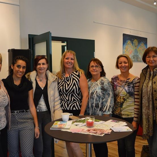 Eine Gruppe von Frauen steht um einen Tisch in einem Raum und lächelt für ein Foto. Der Tisch hat Broschüren und eine Kerze. Sie sind in verschiedene Winterkleidung gekleidet.