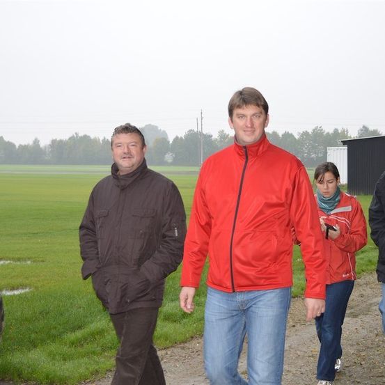 Vier Personen gehen auf einem Feldweg in einem Feld, wobei ein Traktorreifen links sichtbar ist. Der Mann im roten Mantel lächelt in die Kamera.