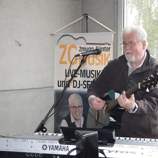 Ein älterer Mann spielt Gitarre und Keyboard vor einem Banner, das 'ZG Musik Live-Musik und DJ-Set' besagt. Er trägt eine braune Jacke und eine Brille. Hinter ihm steht ein Mikrofon auf einem Ständer.
