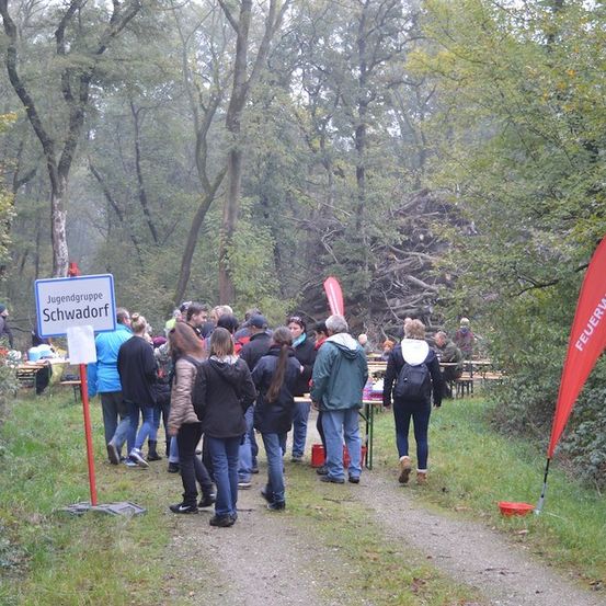 Eine Gruppe von Menschen versammelt sich in einem Wald, einige tragen Jacken und Stiefel. Es gibt Tische und Bänke mit Essen und Getränken aufgestellt. Ein Schild zeigt Jugendgruppe Schwadorf. Ein rotes Banner befindet sich auf der rechten Seite des Bildes.