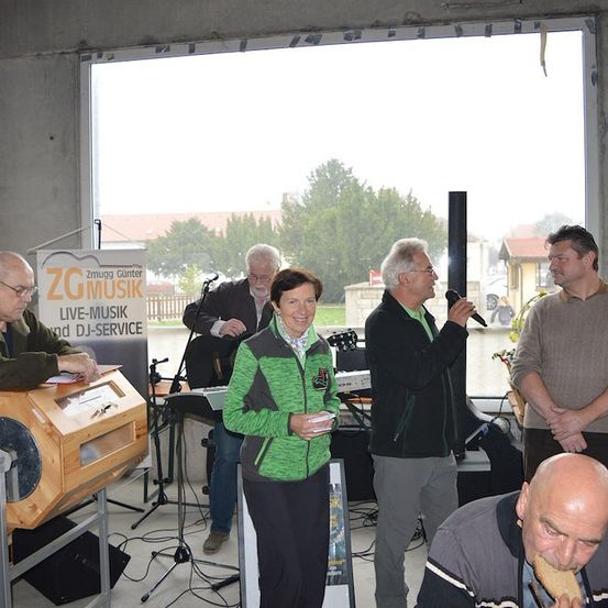 Eine Gruppe von Menschen versammelt sich in einem Raum, möglicherweise für eine musikalische Veranstaltung. Ein Mann in einer grünen Jacke steht vor einem Mikrofon, während ein anderer Mann in einer schwarzen Jacke ein Mikrofon hält. Eine Frau in einer grünen Jacke steht neben einer Holzkiste. Ein Banner mit dem Text 'ZGMUSIK' hängt an der Wand.
