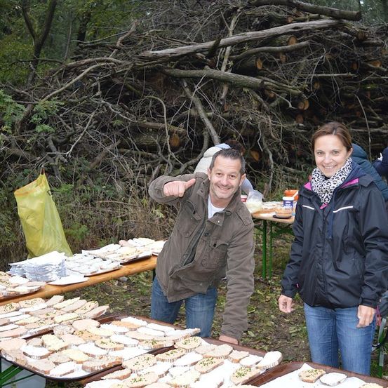 Ein Mann und eine Frau stehen neben einem Tisch mit Essen im Freien. Hinter ihnen ist ein Holzstapel und eine gelbe Tasche zu sehen.