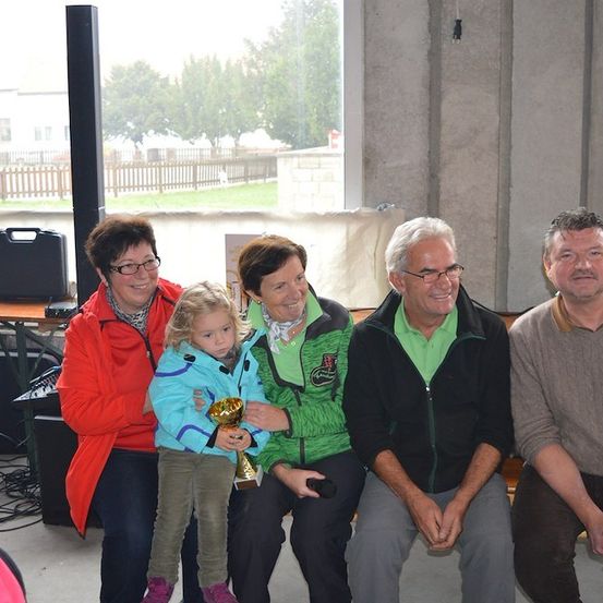 Fünf Erwachsene und ein junges Mädchen posieren für ein Foto in einem Raum mit einem Fenster, das einen Rasen und Bäume zeigt. Das Mädchen hält einen Pokal.