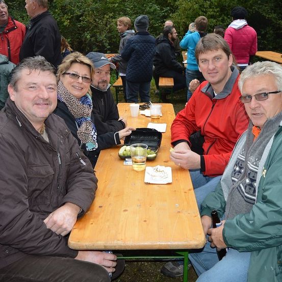 Eine Gruppe von Menschen sitzt an einem Holztisch im Freien, lächelt und posiert für ein Foto. Sie tragen Jacken und einige haben Brillen. Es gibt Tassen und Teller auf dem Tisch, und einige Menschen stehen hinter ihnen.