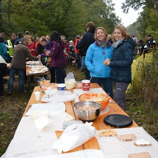 Eine Gruppe von Menschen versammelt sich um einen langen Tisch voller Essen, möglicherweise bei einem Picknick oder einer Outdoor-Veranstaltung. Zwei Frauen stehen vor dem Tisch, eine hält ein Getränk.