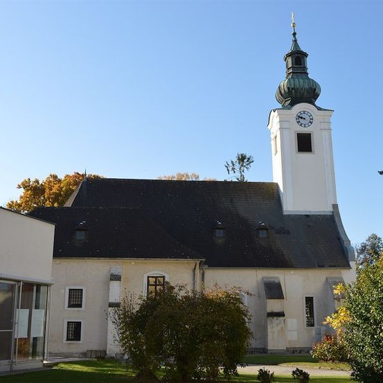 Eine weiße Kirche mit schwarzem Dach und einem hohen Turm mit einer Uhr, umgeben von Bäumen und Pflanzen unter einem klaren blauen Himmel.