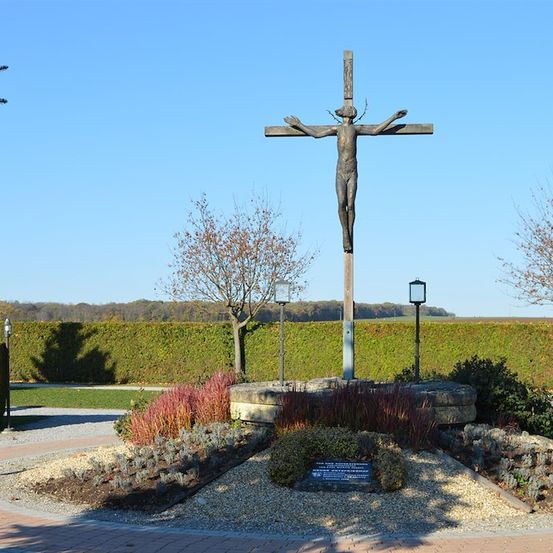 Ein bronzenes Kruzifix steht prominent in einem Garten, umgeben von Grünpflanzen und einem Steinsockel. Das Kreuz befindet sich auf einem gepflasterten Weg mit einem klaren blauen Himmel im Hintergrund.