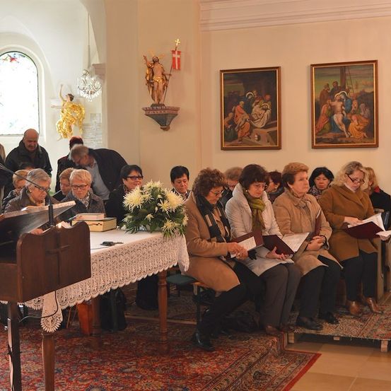 Eine Gruppe von Menschen versammelt sich in einer Kirche, einige sitzen auf Stühlen und andere stehen, alle halten Bücher. Eine Frau liest aus einem Buch an einem Podium.