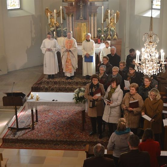 Eine Gruppe von Menschen versammelt sich in einer Kirche, einige stehen und andere sitzen. Die Priester stehen auf dem Altar, halten ein Buch und eine blaue Mappe. Der Altar ist mit Blumen, Kerzen und einer Kristallleuchter geschmückt. Die Kirche hat einen Holzboden und weiße Wände.