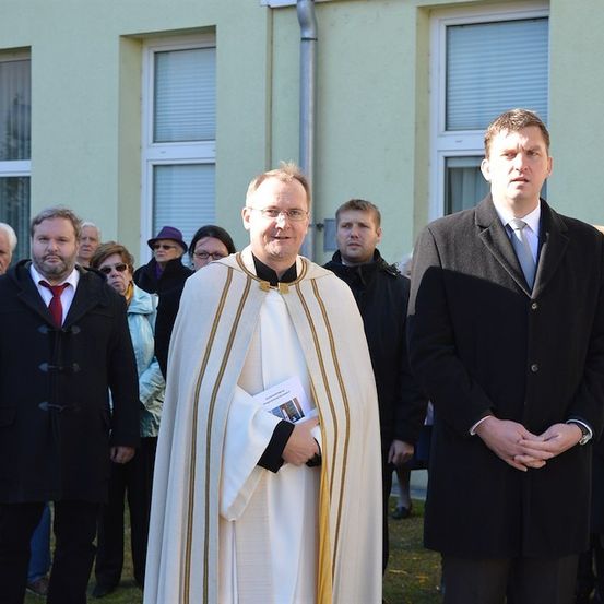 Ein Priester in einer weißen Robe steht neben einem Mann in einem schwarzen Anzug, beide schauen nach vorne. Hinter ihnen stehen mehrere Menschen vor einem Gebäude.