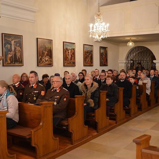 Eine Gruppe von Menschen in Uniform und ziviler Kleidung sitzt auf Kirchenbänken in einer Kirche mit religiösen Gemälden an der Wand und einer von der Decke hängenden Kristallleuchter.