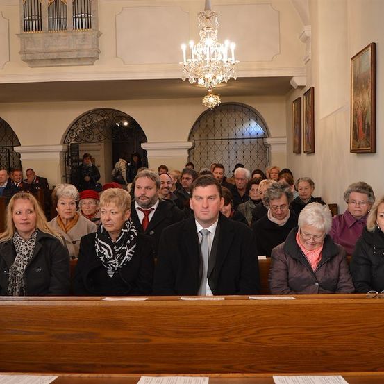 Eine Gruppe von Menschen, sowohl Männer als auch Frauen, sitzen in einer Kirche. Sie sind formell gekleidet und scheinen an einem Gottesdienst teilzunehmen. Der Raum ist gut beleuchtet, und es gibt dekorative Elemente wie Kronleuchter und Gemälde an den Wänden.