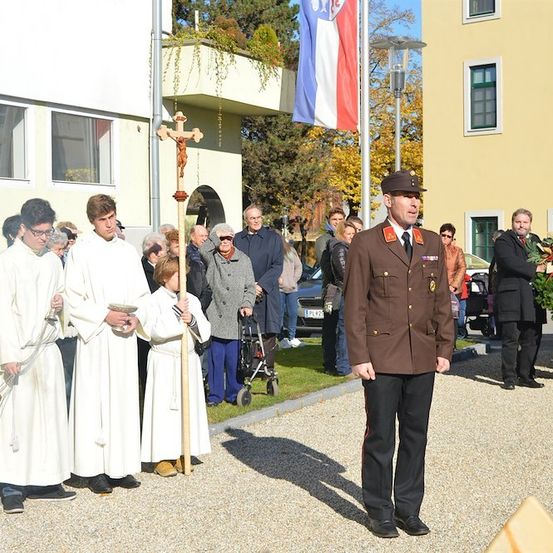 Eine feierliche Zeremonie mit Priestern und einem Militäroffizier, einem Kreuz und einem Fahnenmast im Hintergrund.