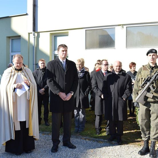 Eine Gruppe von Menschen steht vor einem Gebäude, einige in formeller Kleidung und andere in Militäruniformen, mit einem Priester, der ein Papier hält, und einem Soldaten mit einem Gewehr.